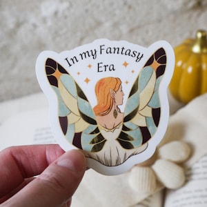 Peut inclure: Autocollant avec l'inscription "In my Fantasy Era" et une illustration d'une femme aux cheveux roux et aux ailes de papillon. Les ailes sont bleues, jaunes et marron. L'autocollant est tenu.