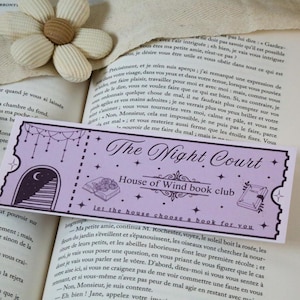 Peut inclure: Un marque-page lavande avec le texte "The Night Court" et "House of Wind book club". Le marque-page présente des illustrations de lune, d'étoiles et de livres. Il repose sur un livre ouvert avec une fleur en tissu.