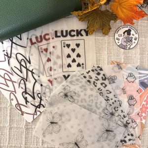 以下が含まれることがあります： さまざまなデザインの装飾的なプランナーインサートのコレクション。トランプのマーク、ハート、蝶、ヒョウ柄のインサートが含まれています。「LUCKY」という言葉が1つのインサートに印刷されています。緑色の革製品が背景に見えます。