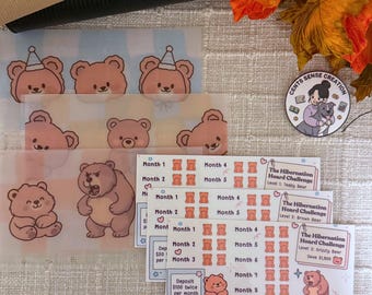May include: A collection of clear plastic budget envelopes featuring cute cartoon bears. The envelopes have different bear designs and text, including "The Hibernation Hoard Challenge" and savings goals. A small round sticker with a girl and a dog is also visible.