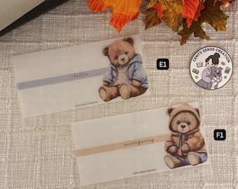 May include: Two white envelopes with teddy bear illustrations. One envelope has the word "bills" in a light purple band, the other says "emergency" in a light brown band. A round sticker with a woman and a dog is also visible.