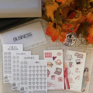 Puede incluir: Un surtido de artículos de papelería, que incluyen sobres de plástico transparente etiquetados como "BUSINESS", y hojas de pegatinas con los textos "I MEAN BUSINESS", "P.O. BOX", "SUPPLIES" y "MISC.". La imagen también presenta pegatinas temáticas "BOSS BABE" y una pegatina con una mujer y un perro.