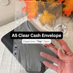 May include: A5 clear cash envelope, zipperless and open at the top. The transparent plastic envelope has pre-punched holes for binder use. The envelope is held over a black fabric wallet.