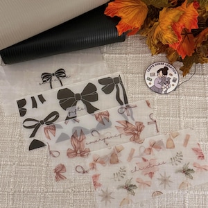 Puede incluir: Sobres de efectivo transparentes de celofán con diseños de lazos y texto. Los sobres están etiquetados como "self care", "gifts", "emergency" y "christmas". Se ve una cartera de cuero negro en el fondo.