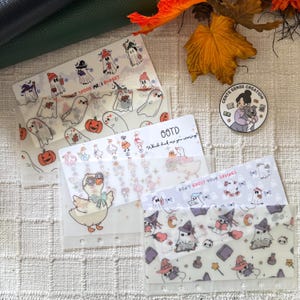 May include: A collection of clear budget envelopes with various designs. One envelope features ghosts and pumpkins, with the text "SPOOK ME, I BUDGET." Another has ducks and the text "OOTD Which duck are you wearing?" A third has ghosts and the text "DON'T GHOST YOUR SAVINGS."