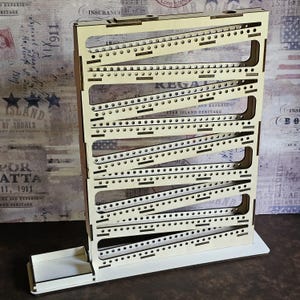 Op de afbeelding: Een houten knikkerbaan spel met een witte achtergrond en zwarte lijnen. Het spel is gemaakt van meerdere niveaus en heeft een klein bakje onderaan om de knikkers op te vangen.