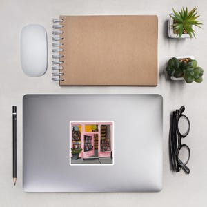 May include: A sticker featuring a digital painting of a pink storefront with a glass door and shelves full of books. The door is open and there is a yellow light shining from inside the store.