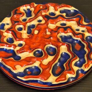 May include: A circular coaster featuring a multi-layered, wavy pattern of orange, blue, and cream-colored wood grain. The surface has a carved, topographical texture with recessed blue areas and raised orange and cream ridges throughout the design.