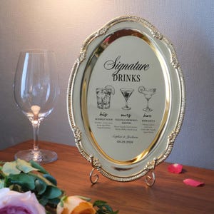 May include: An oval gold-toned metal tray with a decorative beaded rim, displayed on a small stand. The center features black printed text reading Signature DRINKS, his WHISKEY SOUR, ours TEQUILA ESPRESSO MARTINI, her MARGARITA, Sophia & Jackson, 08.25.2026.