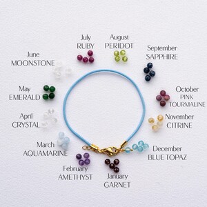 May include: A blue cord bracelet with a gold clasp, encircled by small gemstone clusters representing birthstones. Gemstones include ruby, peridot, sapphire, and topaz. Text labels identify each stone by month.