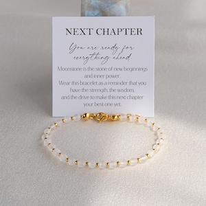 May include: A beaded bracelet featuring small, translucent white moonstone beads interspersed with tiny gold-toned metal spheres. The bracelet includes a gold-toned lobster clasp and is displayed in front of a white card with the text NEXT CHAPTER.
