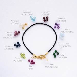 May include: A black cord bracelet with a gold clasp and twelve different coloured gemstone beads. Each bead represents a different month of the year. The beads are arranged in a circle around the bracelet, with the month name and gemstone name written below each bead.