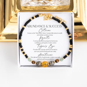 May include: A beaded bracelet featuring small black beads, gold-toned accents, and larger central stones including yellow citrine, metallic pyrite, brown tiger's eye, and green aventurine. The display card reads ABUNDANCE & SUCCESS.
