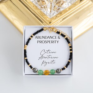 May include: A beaded bracelet featuring small black and gold-tone beads, with three larger central stones in green, yellow, and metallic grey. The bracelet is displayed on a white card printed with the text ABUNDANCE & PROSPERITY and Citrine Aventurine Pyrite.