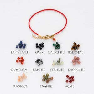 May include: A red string bracelet with a gold clasp and a selection of gemstone beads in various colors, including lapis lazuli, onyx, malachite, tiger's eye, carnelian, hematite, prehnite, rhodonite, sunstone, unakite, and agate.