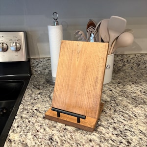 May include: A wooden stand with a black metal handle, designed to hold a tablet or cookbook upright. The stand is made of natural wood with a rustic finish and is sitting on a granite countertop.