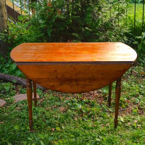 May include: A vintage wooden drop-leaf table with a warm, reddish-brown finish. The table has a rounded top and four slender, turned legs. The table is set outdoors in a grassy area, surrounded by greenery.