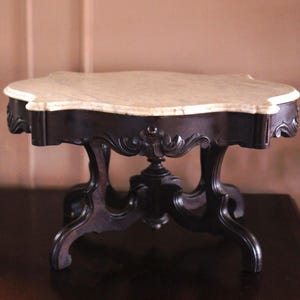 May include: A small, antique table with a marble top and dark wood base. The table has an ornate, carved design on the legs and apron. The marble top is light-colored with a scalloped edge. The table is approximately 30 inches long.