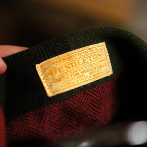 May include: Close-up of a Pendleton label on a red and black patterned wool garment. The label is rectangular with the Pendleton logo and text. The garment has a dark green collar.