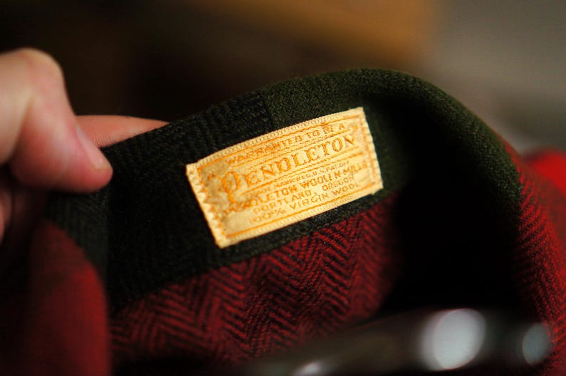 May include: Close-up of a Pendleton label on a red and black patterned wool garment. The label is rectangular with the Pendleton logo and text. The garment has a dark green collar.