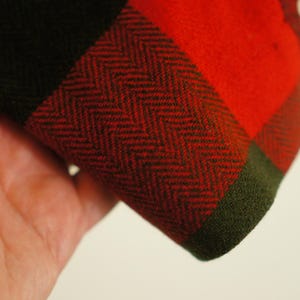 May include: Close-up of a section of fabric with a red and black plaid pattern. The fabric has a herringbone weave in the red and black sections, with a green border. The fabric is held in a hand.