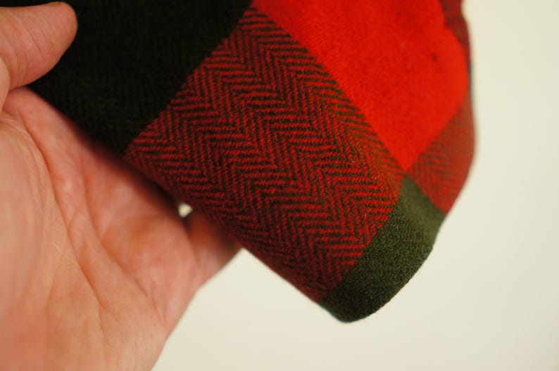 May include: Close-up of a section of fabric with a red and black plaid pattern. The fabric has a herringbone weave in the red and black sections, with a green border. The fabric is held in a hand.