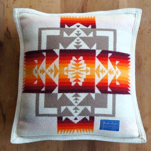 May include: Square decorative pillow with a geometric pattern in shades of red, orange, and brown on a cream background. The pillow has a white border and a blue label that reads "Braver State".