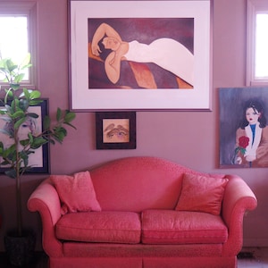 May include: A pink sofa with two matching pillows sits beneath framed artwork in a room with mauve walls. The artwork includes a large print of a reclining figure and two smaller framed pieces. A potted plant is visible on the left.