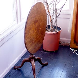 May include: A vintage, oval-shaped wooden table with a dark brown finish. The table has a three-legged base and is positioned near a window. The table's surface shows a star-shaped pattern. The table is a classic piece of furniture.