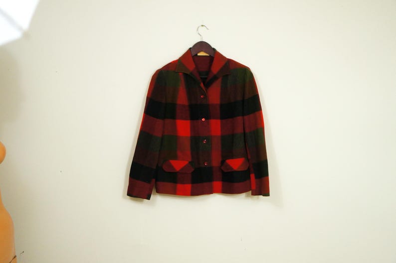 May include: A vintage plaid jacket in red, black, and green hangs on a wooden hanger. The jacket features a collared neckline, button-down front, and two front pockets. The fabric has a textured appearance.
