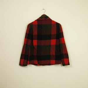 May include: A red, black, and green plaid jacket hangs on a hanger against a white wall. The jacket has a collared neckline and long sleeves. The plaid pattern is a classic design.