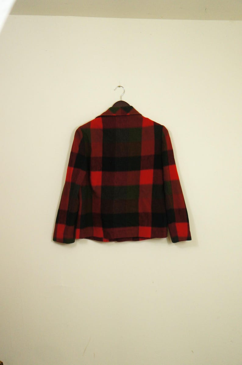 May include: A red, black, and green plaid jacket hangs on a hanger against a white wall. The jacket has a collared neckline and long sleeves. The plaid pattern is a classic design.