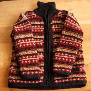 May include: A zip-up knit sweater featuring a high collar and a repeating horizontal pattern in shades of deep red, black, and beige. The garment is constructed with a thick, textured yarn and includes a full-length front zipper closure.