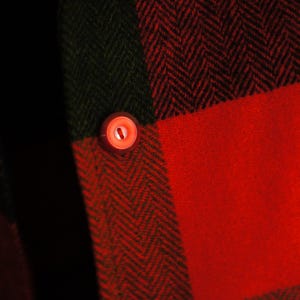 May include: Close-up of a red and black plaid fabric with a herringbone pattern. A small, round, red button is visible on the fabric. The fabric is likely part of a shirt or jacket.