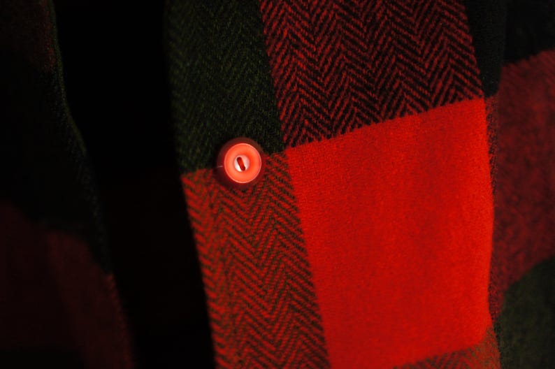 May include: Close-up of a red and black plaid fabric with a herringbone pattern. A small, round, red button is visible on the fabric. The fabric is likely part of a shirt or jacket.