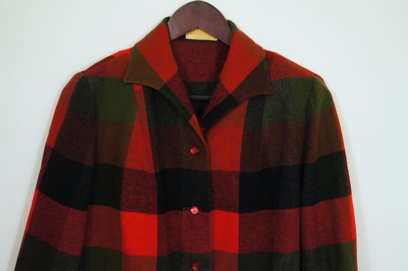 May include: A vintage, button-down coat in a bold plaid pattern of red, black, and olive green. The coat features a classic collar and is displayed on a wooden hanger. The fabric appears to be a warm, wool blend.