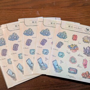 Puede incluir: Cinco hojas de pegatinas idénticas con ilustraciones coloridas de varios cristales y minerales. Cada hoja tiene un encabezado con el texto 鉱石 sticker y muestra múltiples diseños de piedras preciosas en tonos azul, morado, rojo y amarillo, todo dentro de fundas protectoras de plástico transparente.