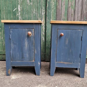 May include: A pair of blue bedside tables with a distressed finish. Each has a single door with a copper-coloured knob and a natural wood top. The tables are set against a weathered green wooden background.