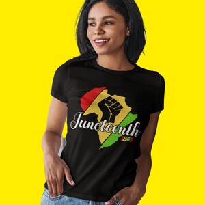 Op de afbeelding: Een zwart T-shirt met een rode, gele en groene afbeelding van een gebalde vuist en de tekst "Juneteenth 1865".
