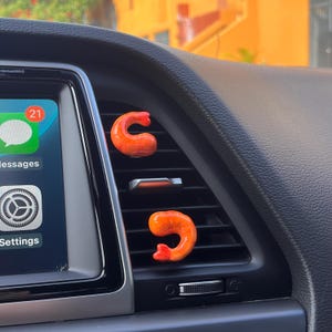 May include: Two orange shrimp-shaped car air fresheners are clipped onto a car vent. The shrimp have a realistic appearance with a curved body and a red tail. The car's dashboard and infotainment screen are visible in the background.