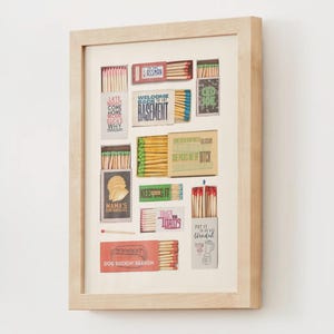 May include: A framed collage of matchboxes with various designs and text, including "Welcome Back to the Basement", "Dog Sniffin' Season", and "Put it on the Blendah".