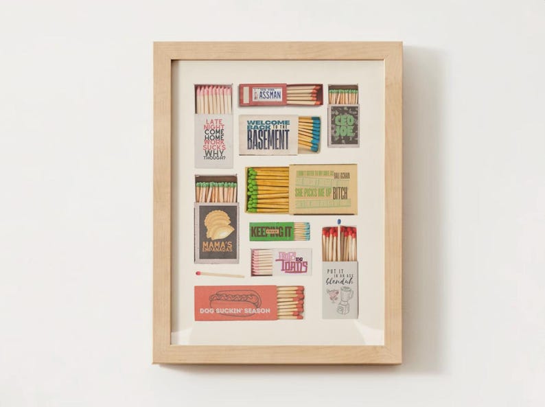 May include: A framed collection of matchboxes with various designs and text, including "Late Night Come Home Work Why Thursday", "Welcome Back to the Basement", "Dog Suckin' Season", and "Put it in a Blender".