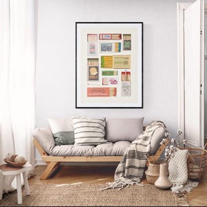 May include: A framed print of a collection of matchboxes with various designs and text, including "Basement", "The Palace of Bitch", and "You Are My Sunshine". The print is displayed on a wall above a light gray futon with a striped throw blanket.