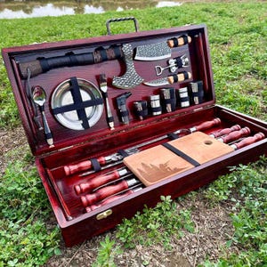Puede incluir: Un juego de picnic de madera rojo oscuro con un hacha, cuchillos, brochetas y una pequeña tabla de cortar. El juego se presenta en una caja de madera abierta con un asa, sobre hierba verde. Incluye plato, tenedor y cuchara.