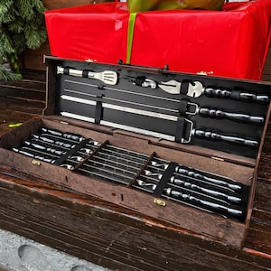 May include: A dark brown wooden barbecue tool set, containing stainless steel skewers and utensils. The set includes a fork, spatula, and skewers, all neatly arranged in an open wooden case. The case is open, revealing the tools inside.