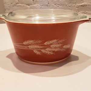 May include: A vintage, rust-colored Pyrex casserole dish with a clear glass lid. The dish features a wheat pattern in white. The dish has handles on either side. A classic piece of kitchenware.