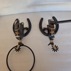 Pode incluir: Dois porta-toalhas de metal preto em forma de ferradura com detalhes de esporas e estrelas. Um tem um anel de toalha circular, o outro uma barra reta. Os suportes são montados em uma parede branca.