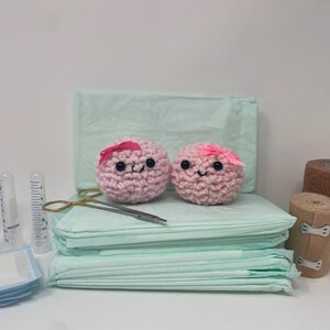 May include: Two pink crocheted plushies with smiling faces and pink bows, alongside medical supplies. The scene includes syringes, gauze pads, medical tape, and scissors, all arranged on a white surface. The plushies are the focal point.