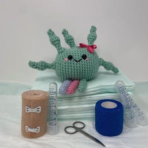 May include: A crocheted teal creature with a pink bow, on blue pads. Medical supplies include a tan bandage roll, blue tape, clear graduated cylinders, and scissors. The creature has a smiling face. Measurements are in cm and grams.