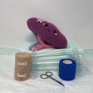 May include: A purple crocheted kidney with a smiling face and a pink flower on top. The kidney is sitting on a stack of white medical pads. There are also bandages, gauze, and medical supplies on the table.